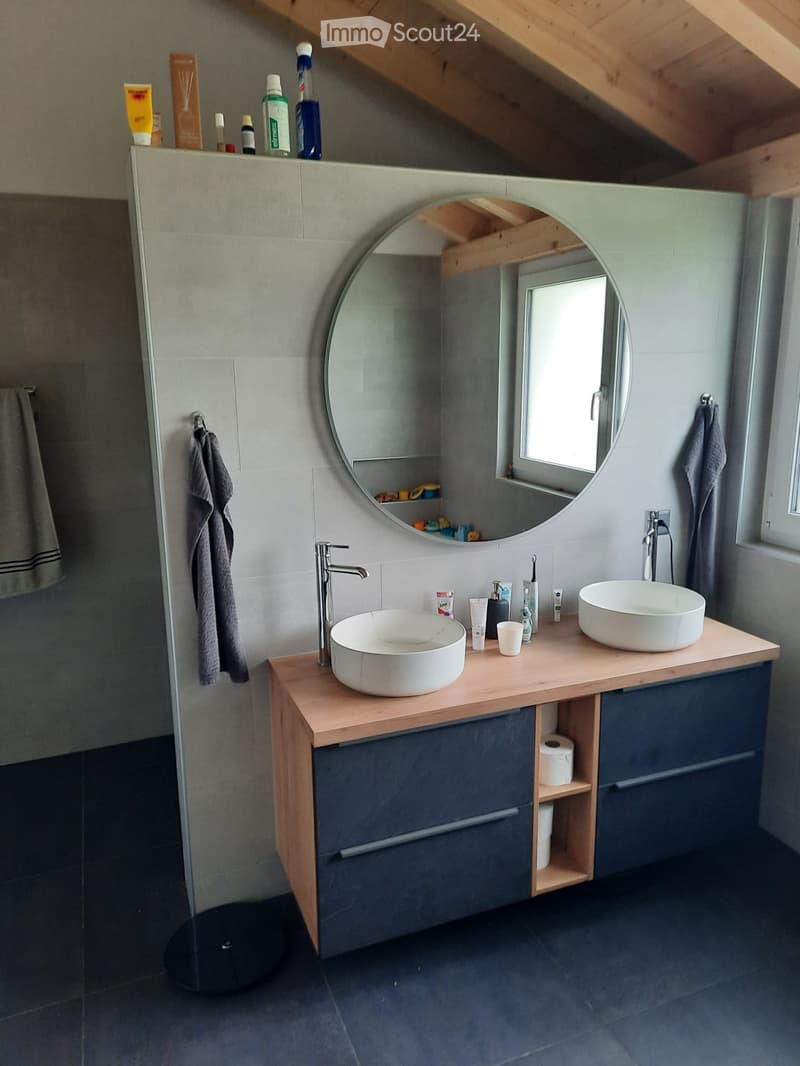 Modern bathroom with two sinks, a large round mirror, wooden vanity, and dark grey tiles.