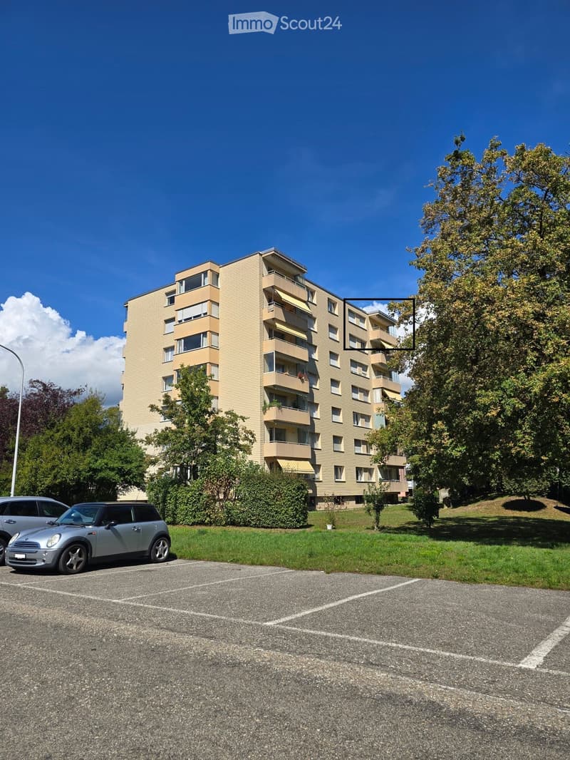 Multi-story residential building with balconies, surrounded by greenery and parked cars.