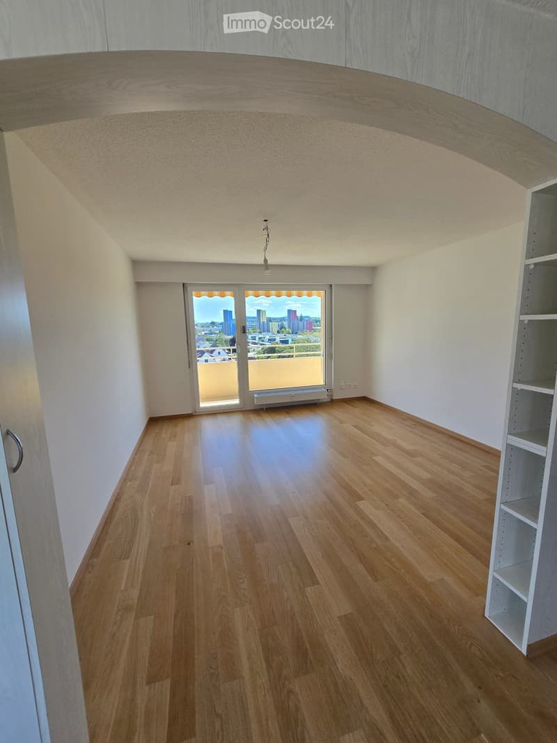 Empty room with wooden flooring, white walls, and a large window offering a city view. The room has a modern, minimalist design with a built-in shelf unit on one side.
