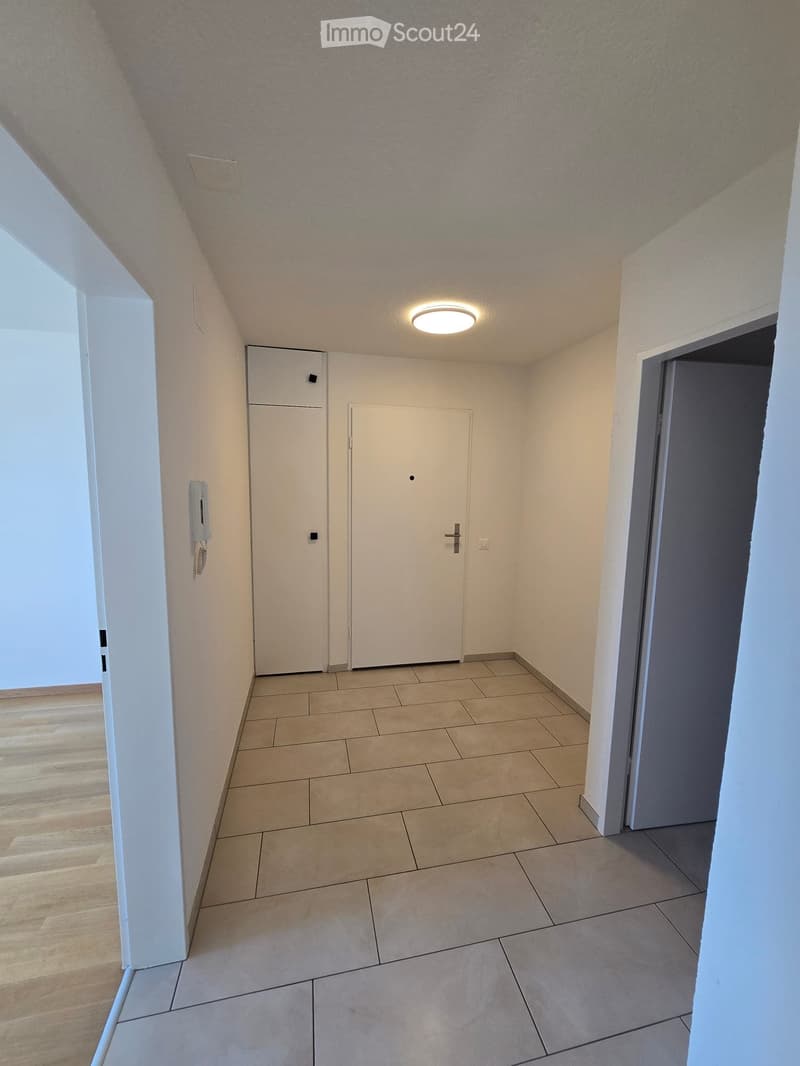 Empty hallway with tiled floor, wooden flooring on one side, white walls, and recessed lighting. Features two doors and a telephone on the wall.