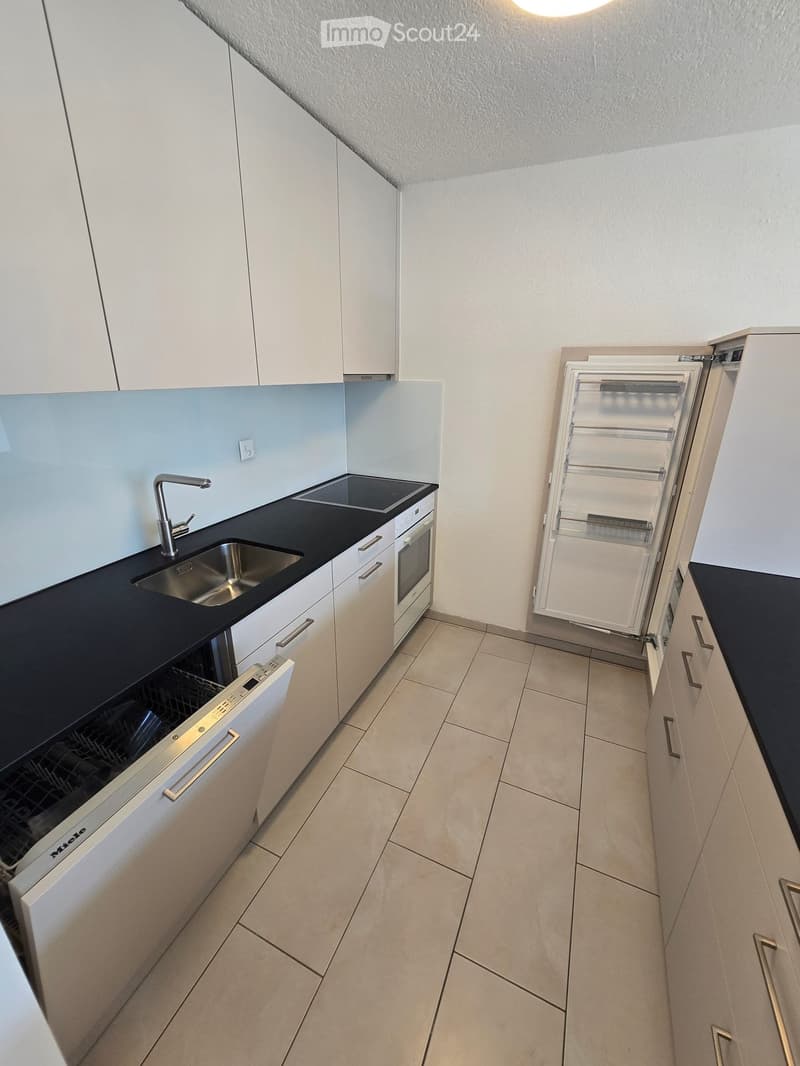 Modern kitchen with white cabinets, black countertops, stainless steel sink, faucet, oven, dishwasher, and open refrigerator.