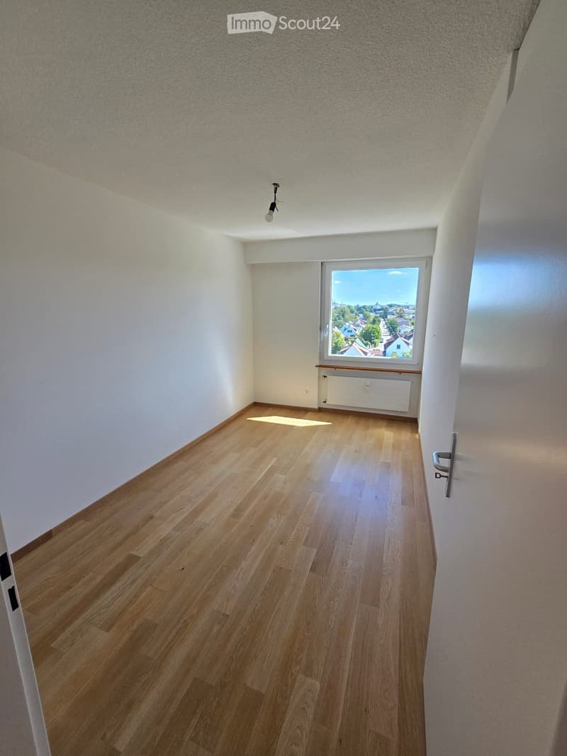 Empty room with wooden flooring, white walls, and a large window showing a view of the outside. The room has a simple, modern design with a single light fixture on the ceiling.