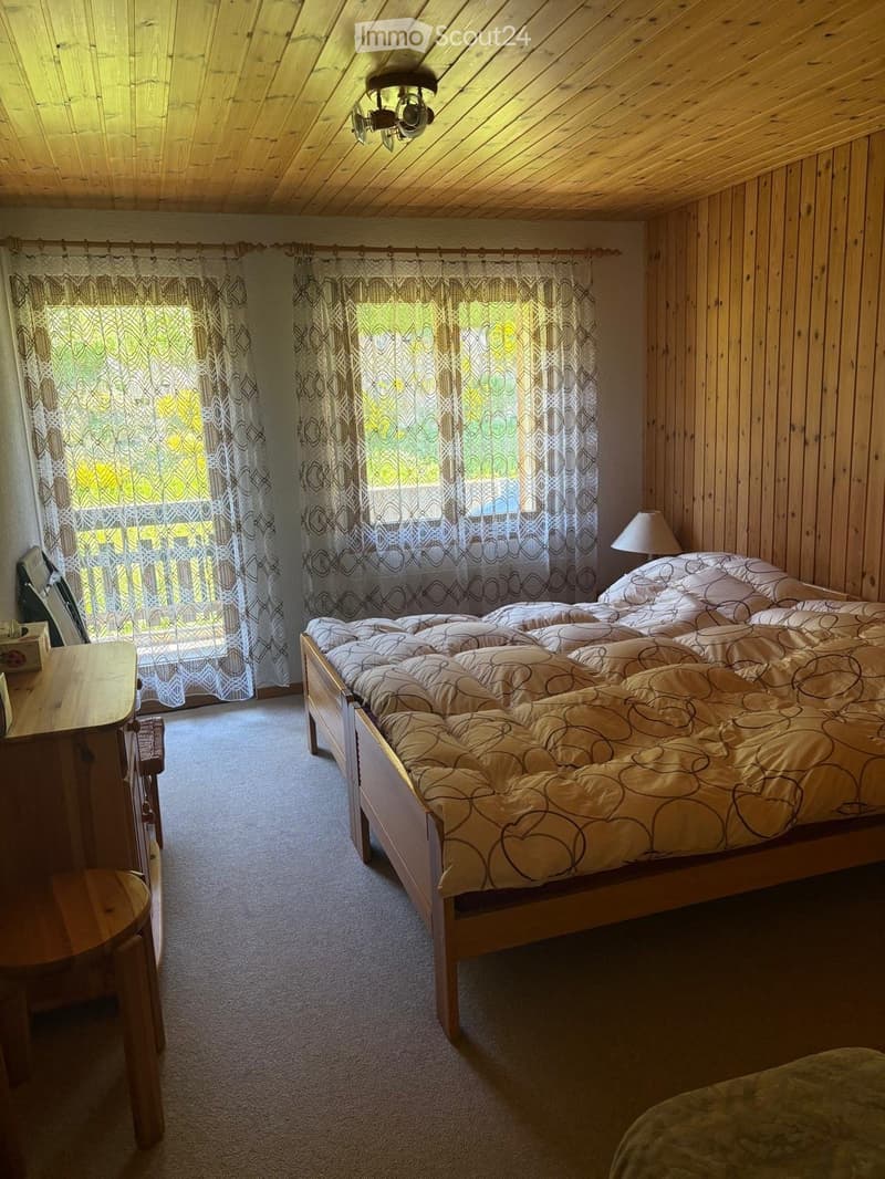 Cozy bedroom with wooden walls and ceiling, large window with patterned curtains, neatly made bed with patterned duvet, nightstand with lamp, and small wooden stool.