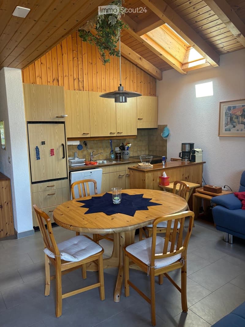 A cozy, well-furnished kitchen and dining area with wooden cabinets, a round wooden dining table with four chairs, a refrigerator, a dishwasher, a sink, and a coffee maker on the counter. The room features exposed wooden beams and a hanging light fixture.