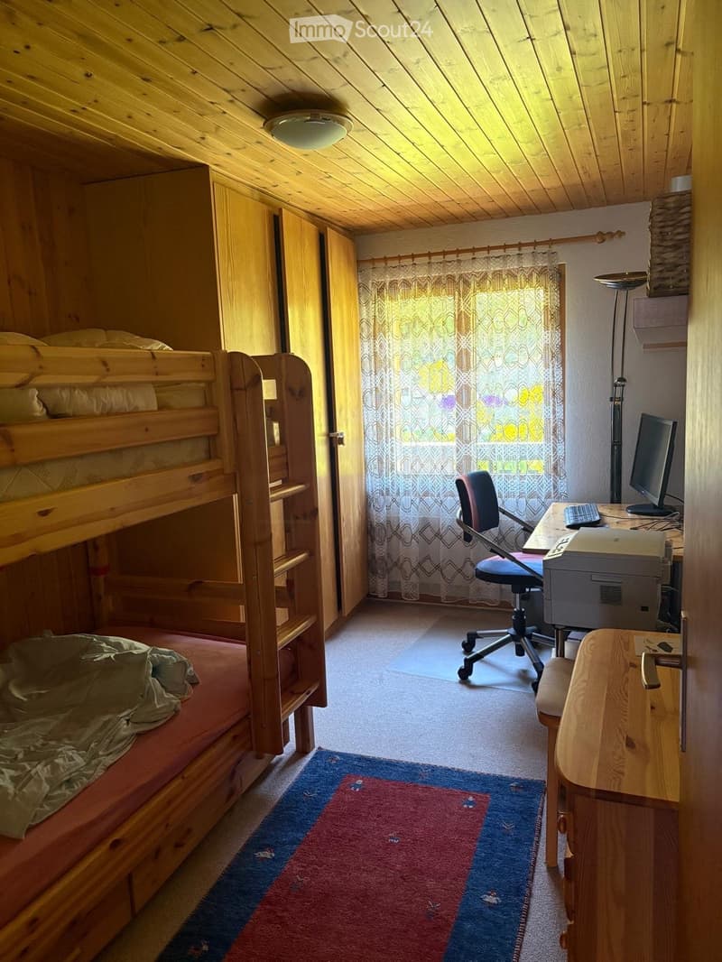 A cozy attic bedroom with a wooden bunk bed, a desk with a computer and printer, a chair, and a colorful rug on the floor.