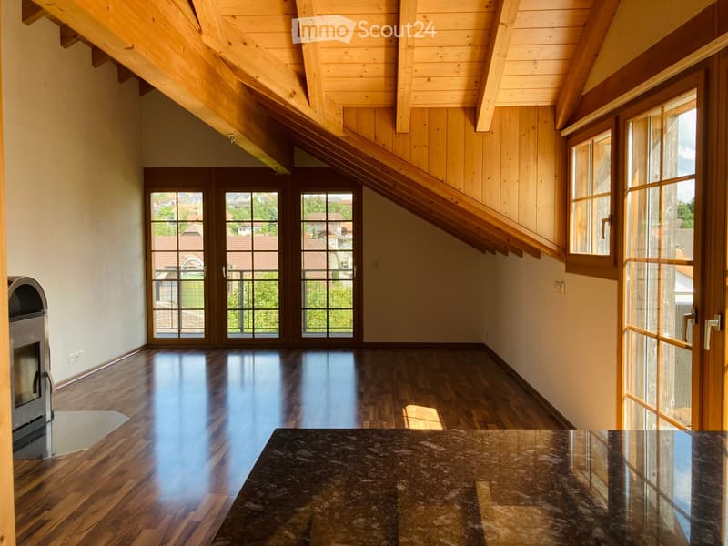 Bright, open loft space with wooden floors, exposed wooden beams, large windows, and a staircase leading to an upper level.