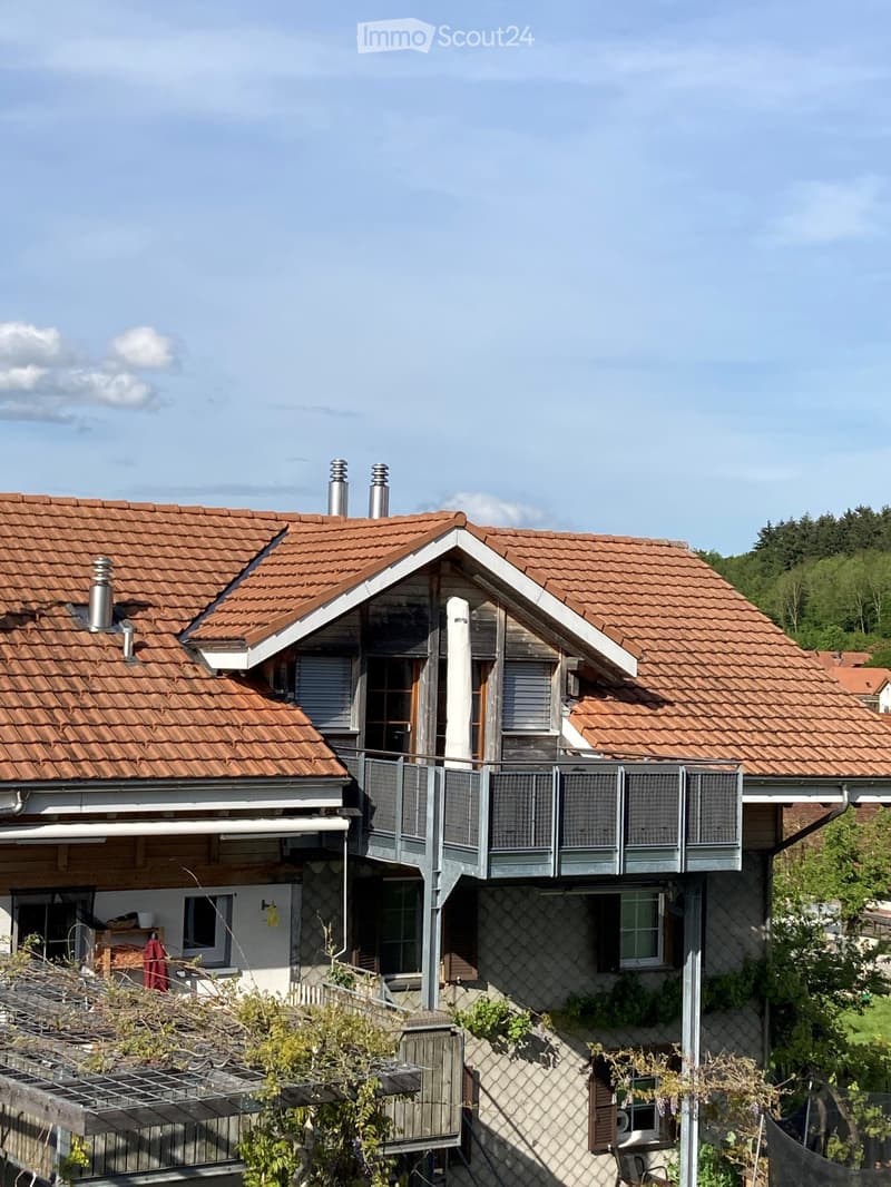 Two-story house with red-tiled roof, multiple chimneys, and a balcony on the upper level. The house features a mix of stone and wooden exterior walls, with several windows and a small garden area in front.