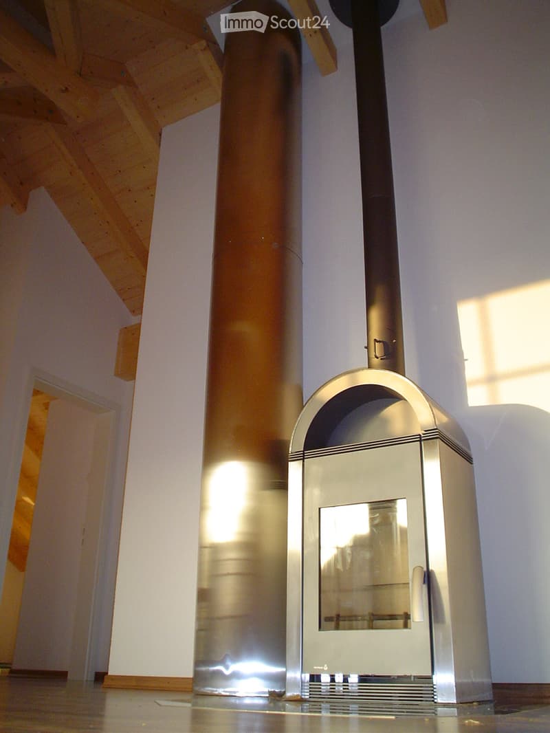 Modern stainless steel fireplace with glass door and chimney, set against white walls and wooden ceiling beams.