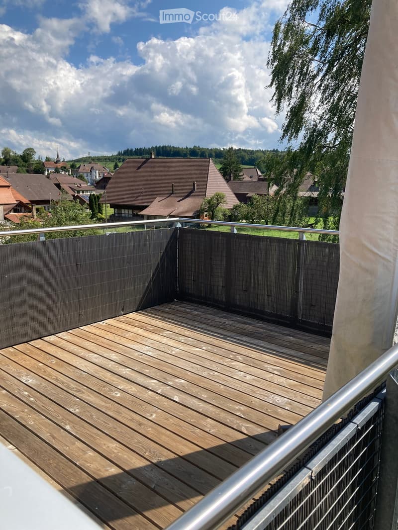 Wooden deck with metal railing, overlooking a residential area with houses and trees under a partly cloudy sky.