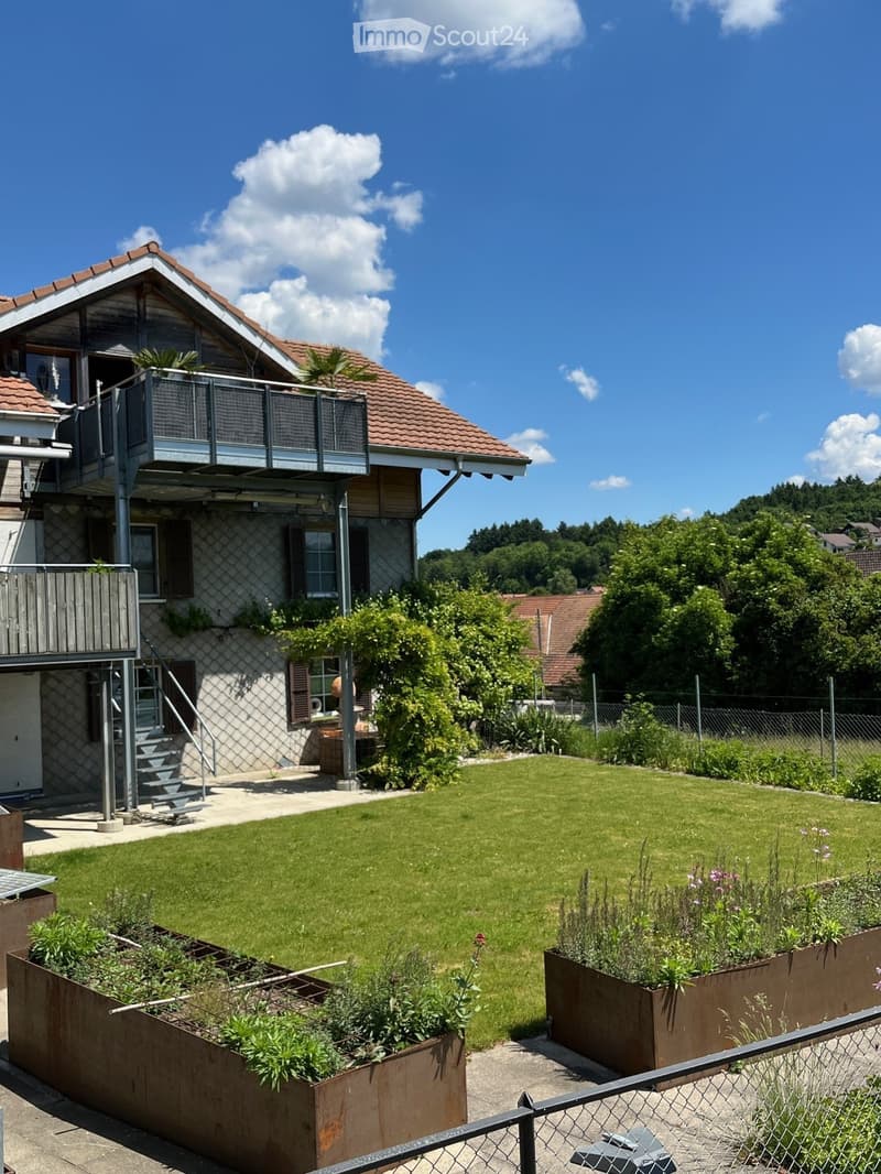 A well-maintained garden with a lawn, raised garden beds, and a modern two-story house with a balcony and staircase leading to the garden.