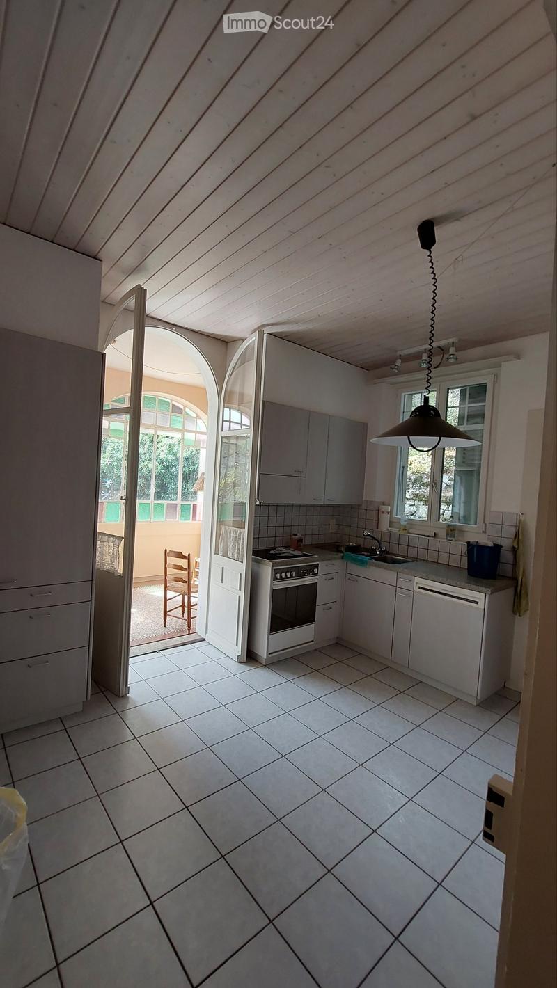 Bright kitchen with white tiled floor, white cabinets, built-in oven, sink, and a hanging light fixture. An arched doorway leads to a balcony area with a wooden chair visible.