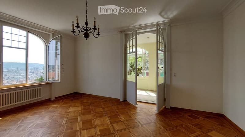 Empty room with wooden parquet flooring, large arched windows offering a city view, and an open doorway leading to another space.