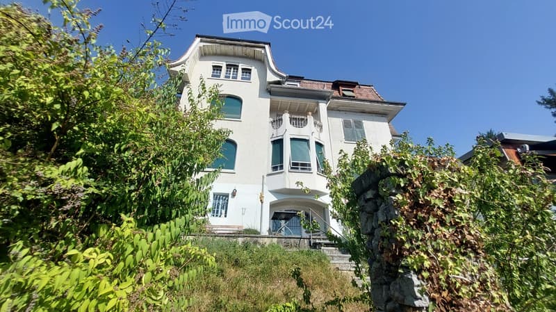 A two-story white house with multiple windows, a balcony, and a staircase leading to the entrance, surrounded by greenery.