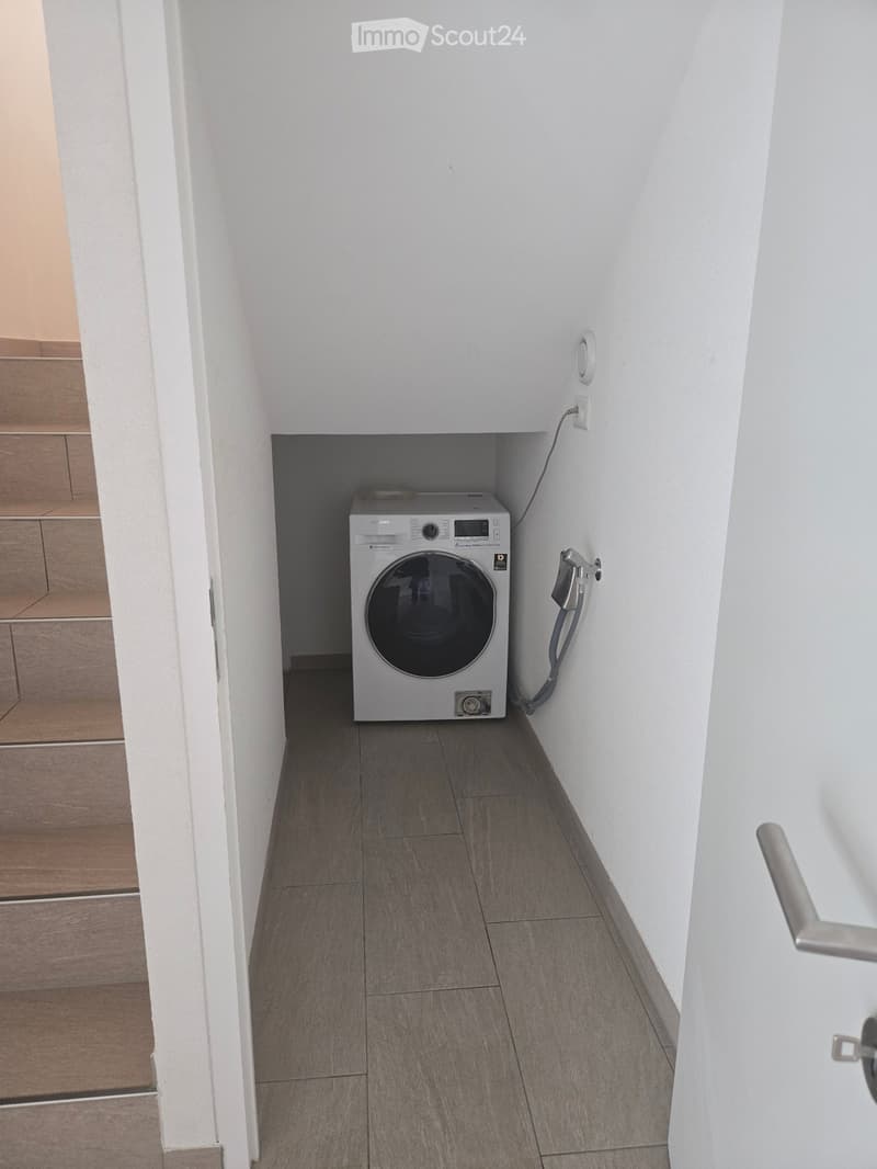 Narrow hallway with a private washing machine placed in a corner, white walls, grey tiled floor, and a visible electrical outlet.