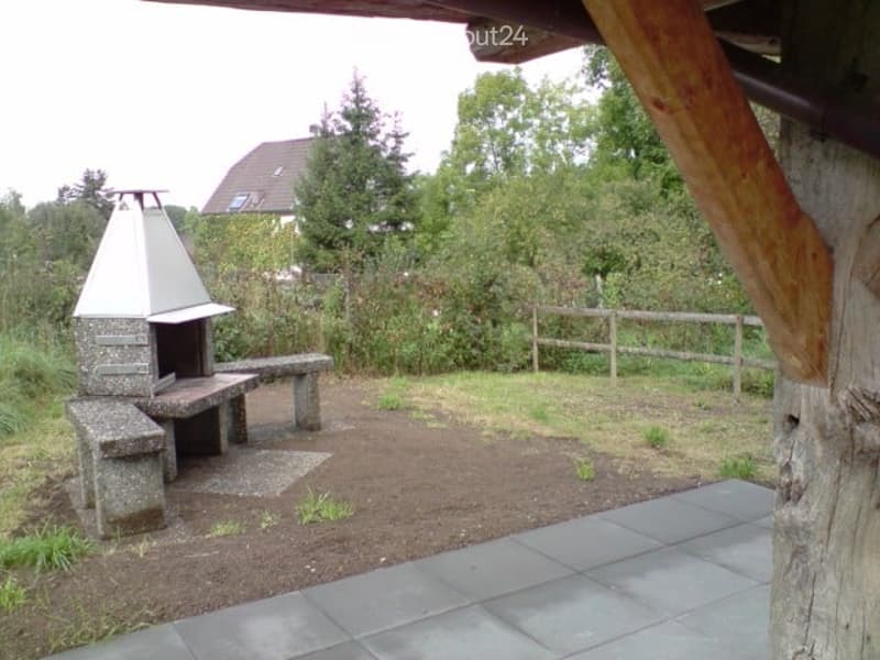Outdoor patio area with a stone barbecue, seating benches, and a wooden fence, surrounded by greenery and a house in the background.
