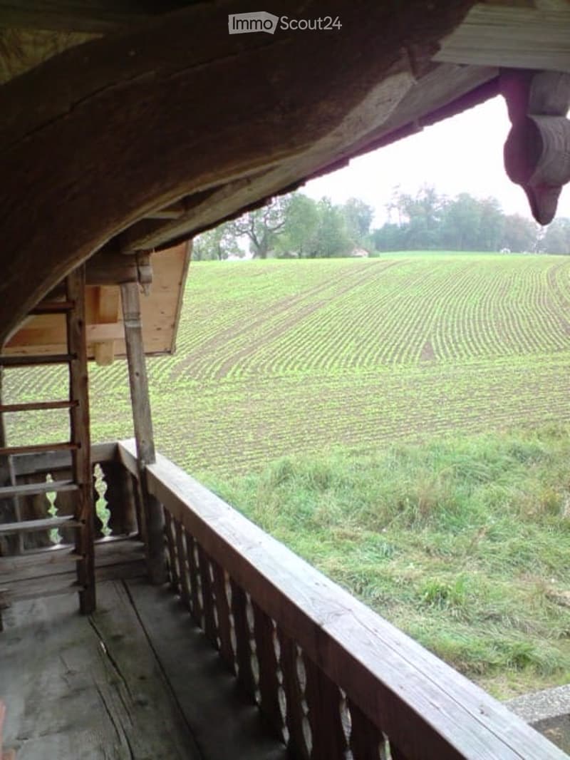 Wooden balcony overlooking a vast, cultivated field with visible rows of crops. The balcony features a wooden railing and a staircase leading up to it.