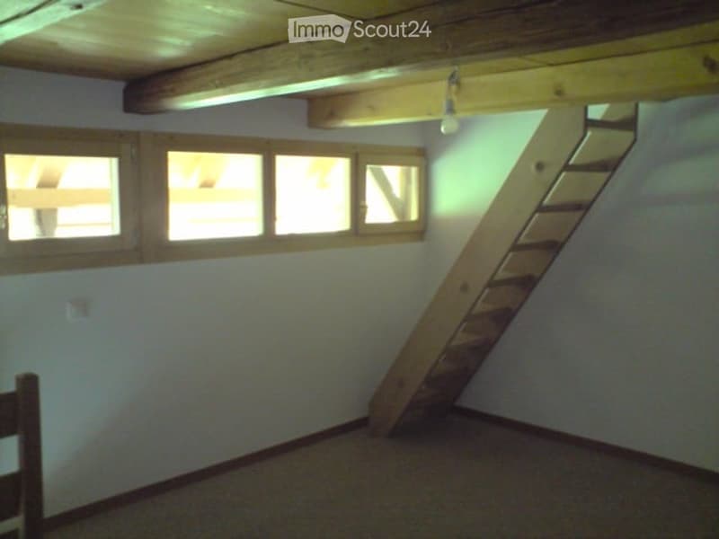 An attic space with exposed wooden beams, a wooden staircase leading up, and several windows letting in natural light. The room is empty with no furniture or furnishings.