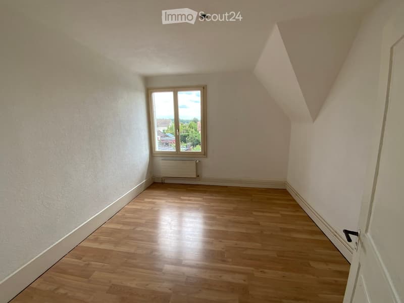 Empty attic room with wooden flooring, white walls, and a single window offering a view of the outside. The room features a slanted ceiling and a radiator below the window.
