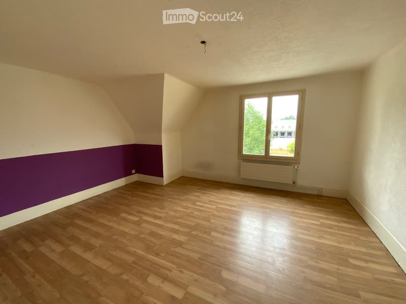 Empty room with wooden flooring, white walls, and a purple accent wall. Features a large window with a view of greenery and a radiator below it.