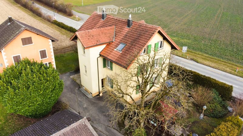 Aerial view of a two-story house with a red-tiled roof, green shutters, and a driveway leading to the entrance. The house is surrounded by a well-maintained garden and is situated near a road with open fields in the background.