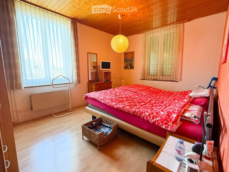 Cozy bedroom with wooden floors, a bed with red and pink bedding, a nightstand, a mirror, a screen, a wicker basket, and a small table with a bottle and lamp.