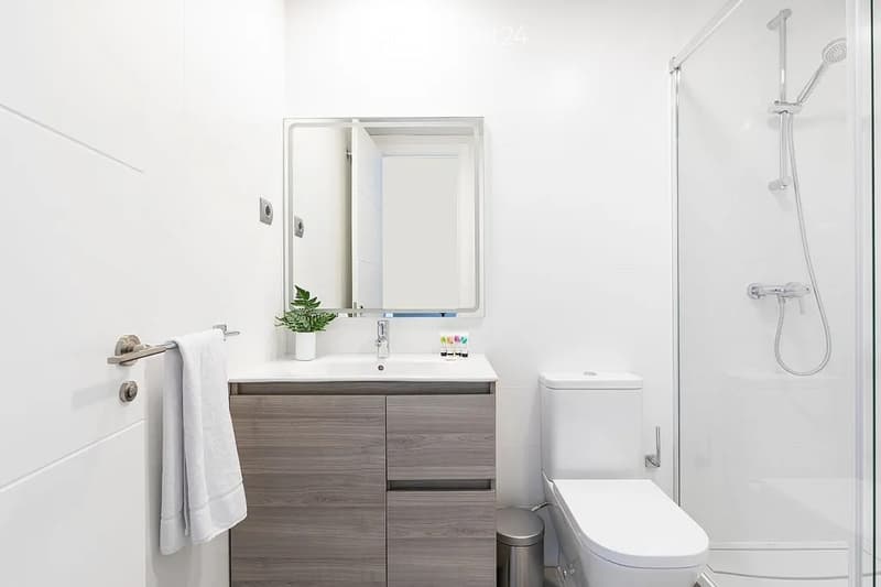 Modern bathroom with a white sink, mirror, toilet, and glass-enclosed shower. Features a wooden cabinet, a towel, and a small plant on the sink counter.