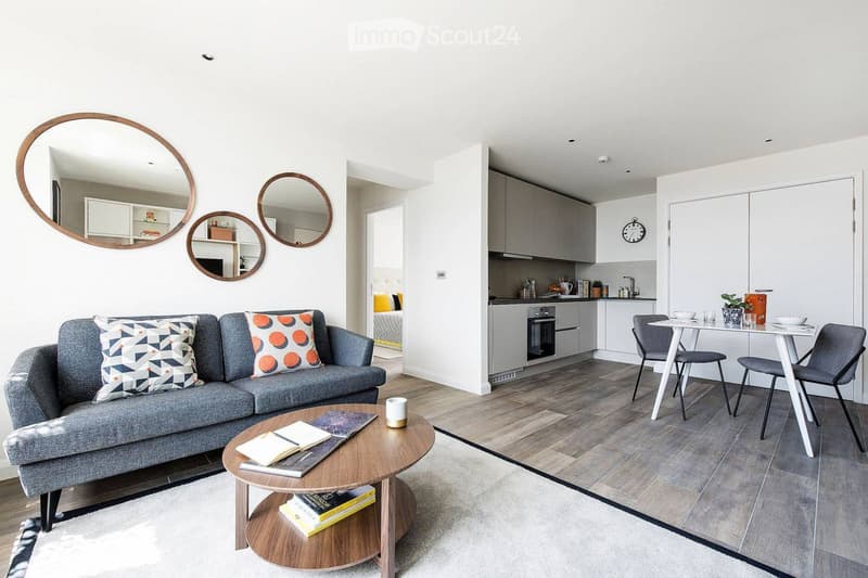 Modern open-plan living and dining area with grey sofa, coffee table, books, and a kitchen with cabinets, oven, and dining table.