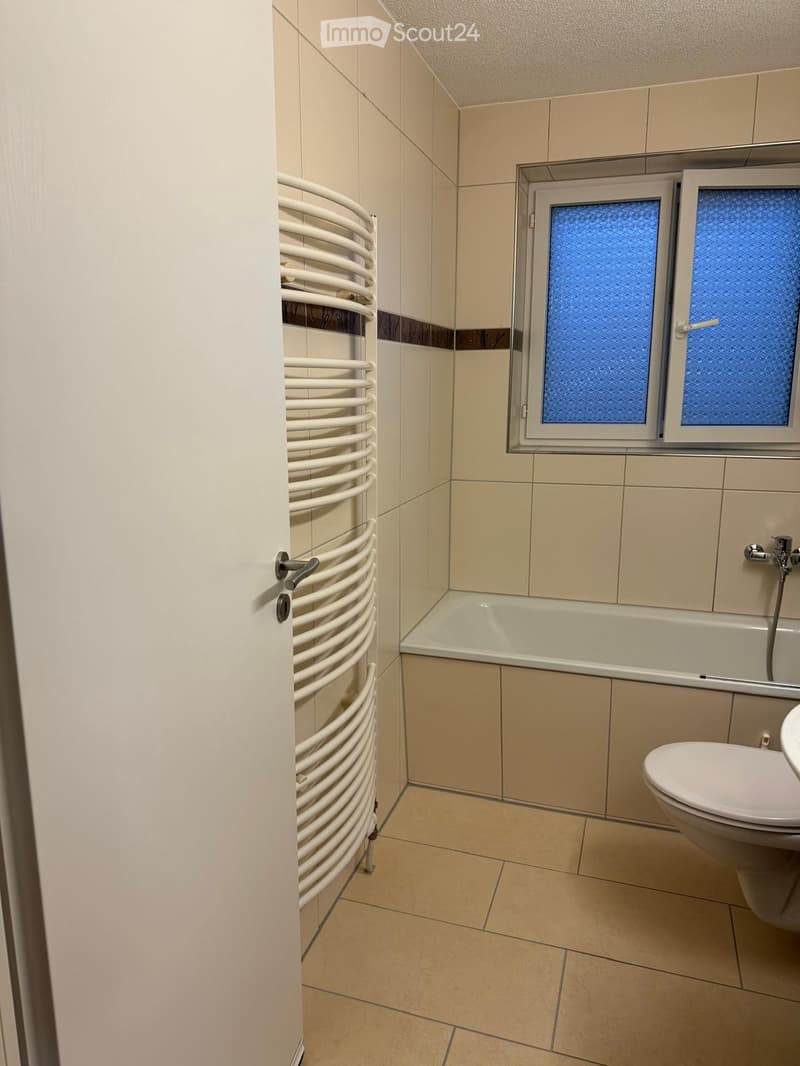 Modern bathroom with beige tiles, a bathtub, toilet, and a white radiator on the wall.