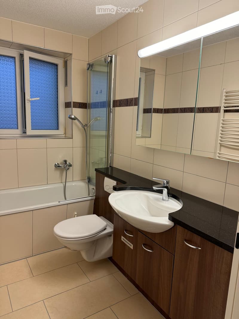 Modern bathroom with beige tiles, a bathtub, toilet, sink, and a glass-enclosed shower area. Features a large mirror with built-in lighting and a window with frosted glass.