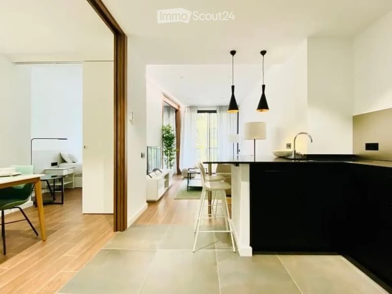 Modern open-plan living and dining area with a sleek kitchen island, dining table, and comfortable seating. The space features light wood flooring, white walls, and contemporary lighting.