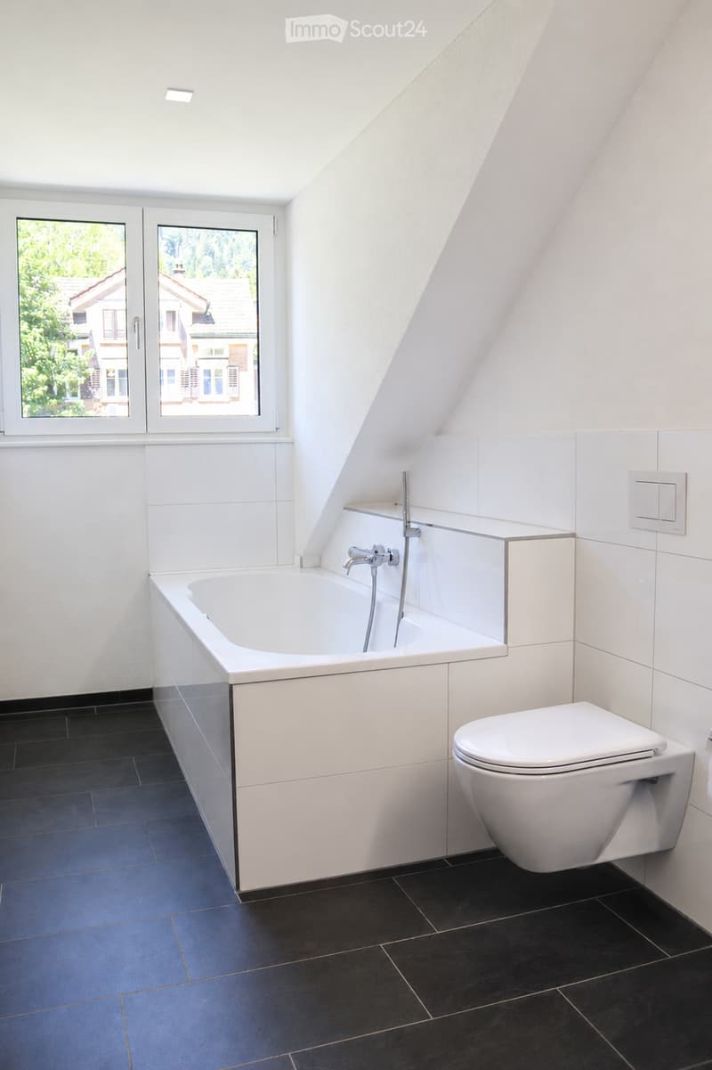Modern bathroom with a white bathtub, toilet, and sleek faucet, featuring dark tiled flooring and white tiled walls, with large windows offering a view of the outside.
