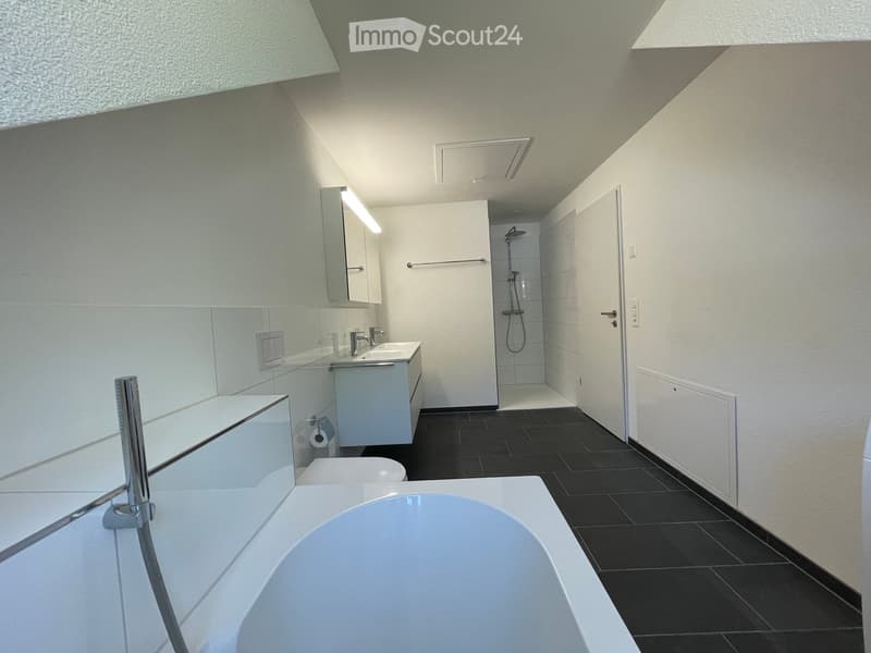 Modern bathroom with a bathtub, sink, toilet, mirror, and a separate shower area with a glass enclosure. The space features sleek white fixtures and black tiled flooring.