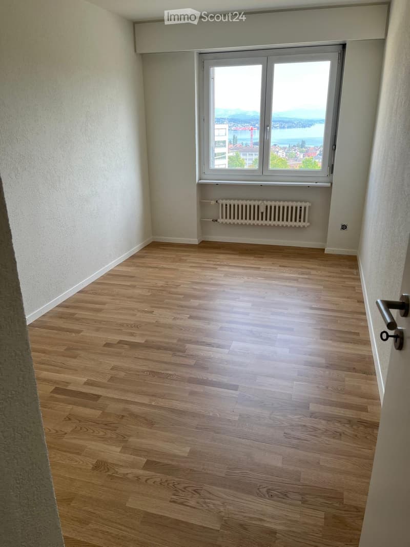 Empty room with light wooden flooring, white walls, and a large window offering a scenic view of a lake and mountains.