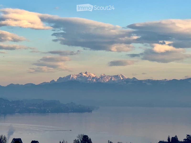 Panoramic view of a lake surrounded by mountains under a partly cloudy sky, with a few houses visible on the shore.