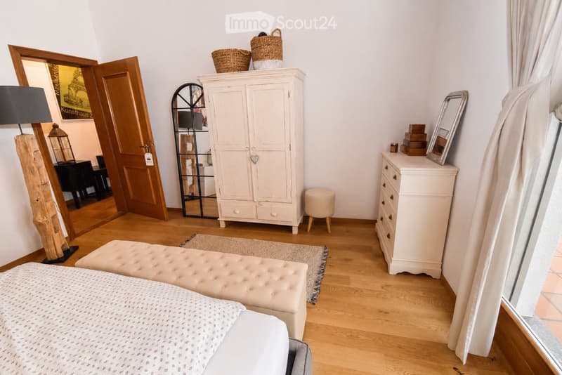 Cozy bedroom with wooden flooring, a bed with patterned bedding, a tufted bench, a white cabinet, a floor lamp, and a mirror on the wall.