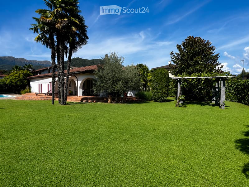 A well-maintained garden with a large, neatly trimmed lawn, a swimming pool, a tall palm tree, and a white house with a red-tiled roof in the background.