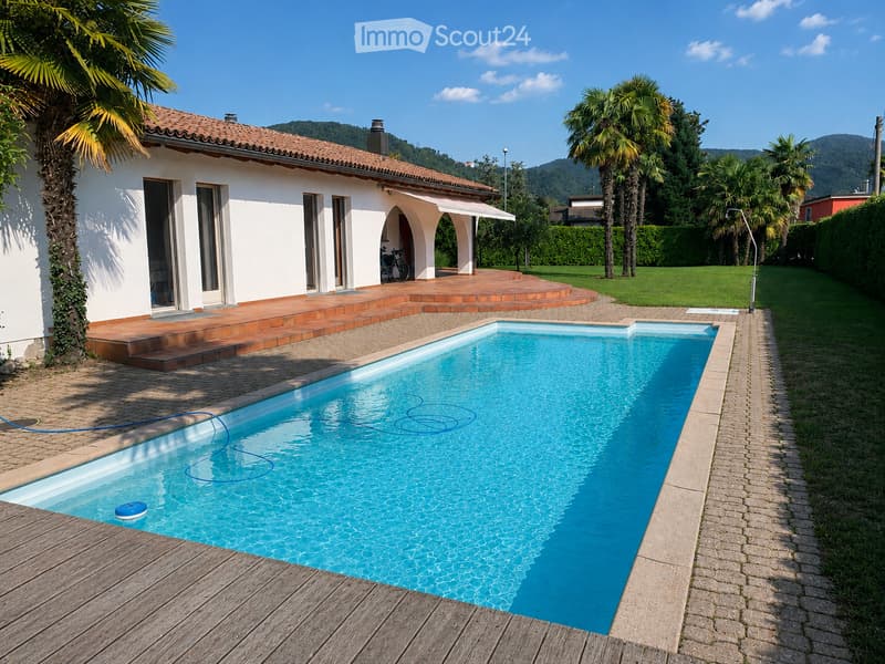 A private garden area with a swimming pool, surrounded by a brick-paved deck, palm trees, and a white house with a tiled roof in the background.