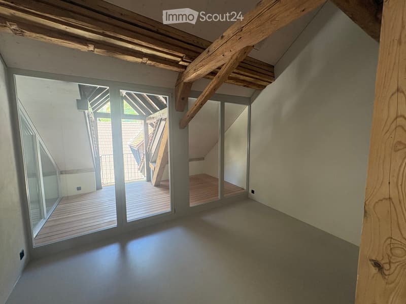Empty attic space with exposed wooden beams, large glass doors leading to a wooden deck, and a smooth concrete floor.