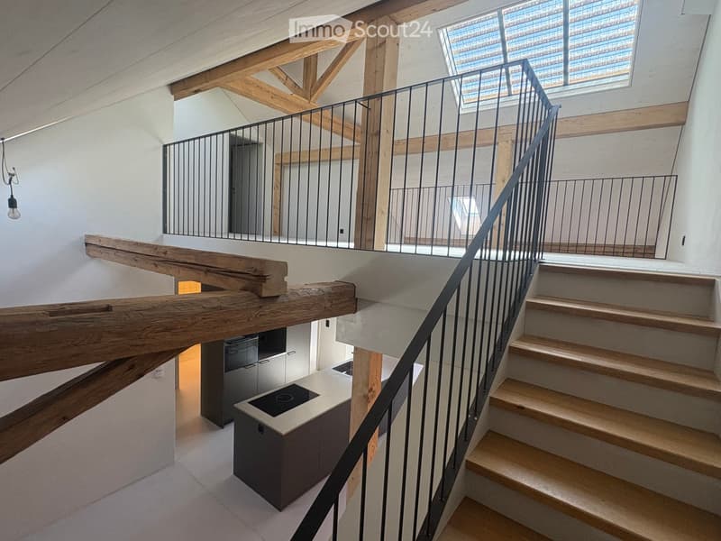 Modern interior staircase with wooden steps, metal railing, and exposed wooden beams. The lower level shows a small kitchen area with a countertop and built-in appliances.