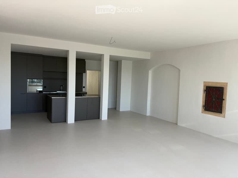 Empty room with white walls, grey cabinets, and a grey floor. The room features an open layout with visible kitchen cabinets and a sink.