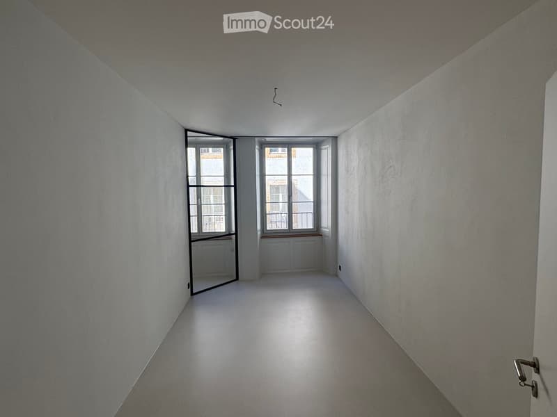 Empty hallway with white walls, a polished floor, and large windows at the end, allowing natural light to enter.