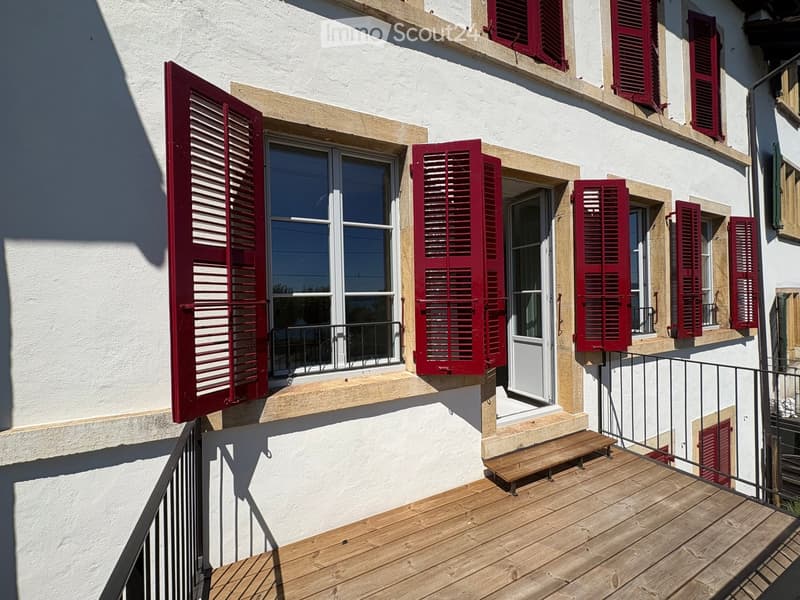 Balcony with wooden decking, metal railing, and a small wooden bench. The building has white walls, red shutters, and multiple windows.