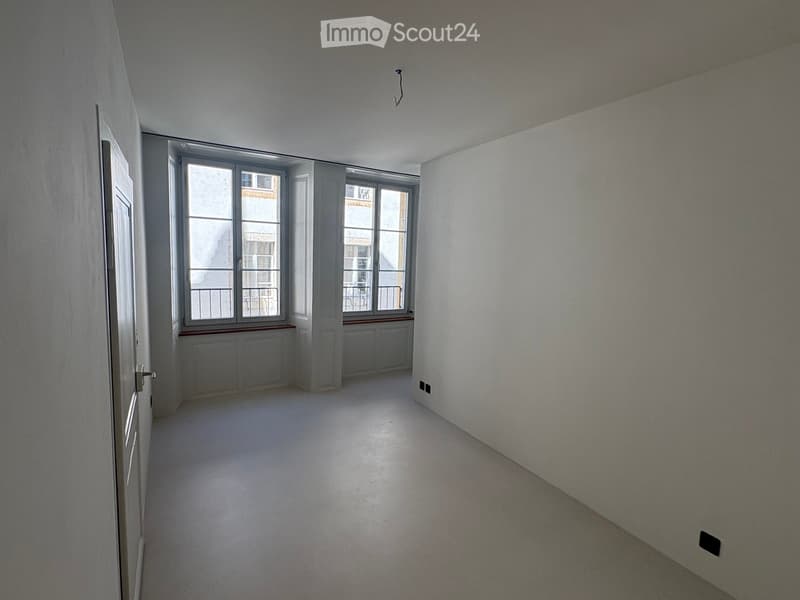 Empty room with white walls, grey carpeted floor, large windows with metal railings, and a door leading to another space.