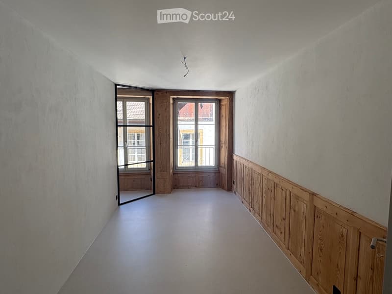 Empty hallway with white walls, wooden paneling on one side, and two windows at the end showing a view of neighboring buildings.