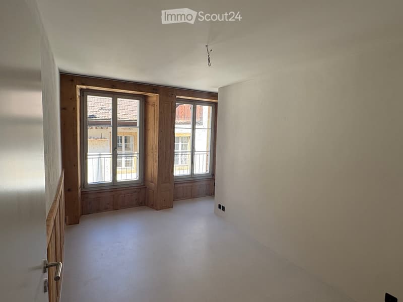 Empty room with light-colored walls, wooden trim around windows, and a carpeted floor. Windows offer a view of a tiled roof and neighboring building.