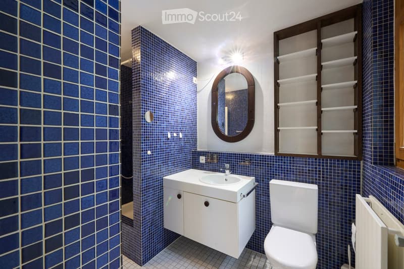 Bathroom with blue tiled walls and floor, white sink, toilet, mirror, and wooden shelves.