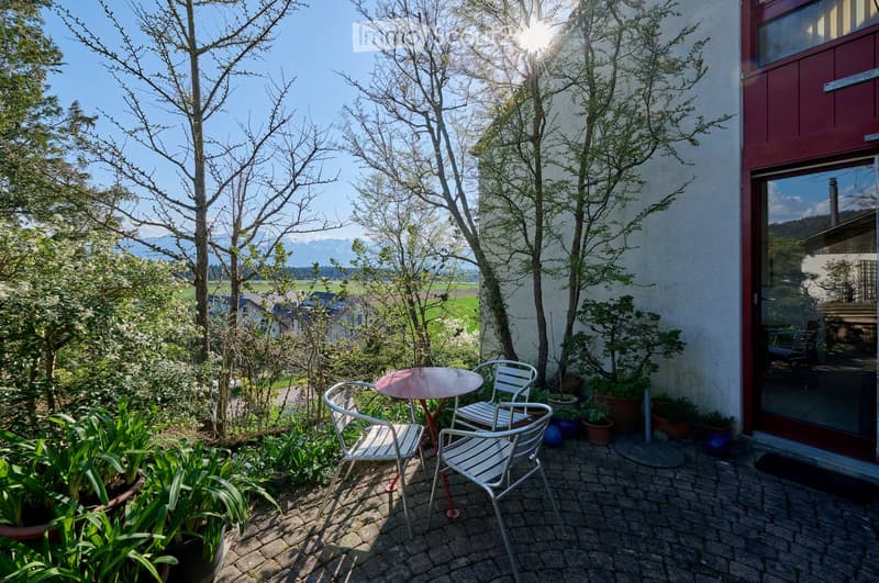 Balcony with a small round table and four chairs, surrounded by potted plants and trees, overlooking a scenic landscape with mountains in the background.