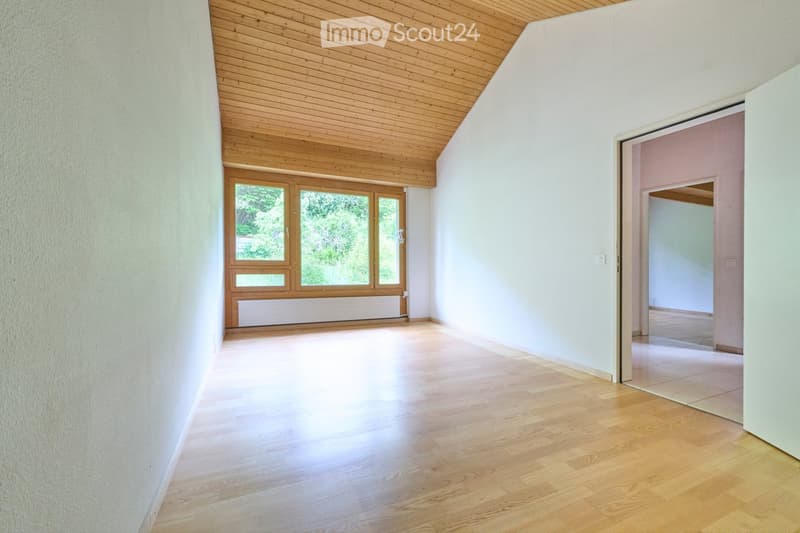 Empty room with light wooden flooring, white walls, and a wooden ceiling. Features a large window with wooden frame and a door leading to another room.