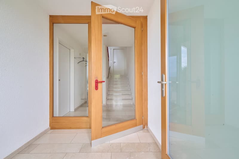 Open wooden door leading to a hallway with tiled flooring and a staircase visible in the background.