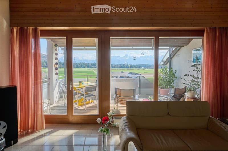 Bright living room with large windows and a balcony overlooking a scenic mountain view. Features a beige sofa, a small table with chairs, and potted plants.